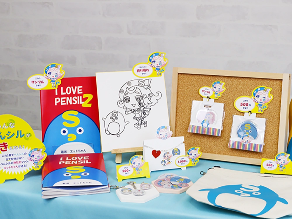 キャラクターグッズの展示。書籍、缶バッジ、キーホルダー、トートバッグなどをテーブルに陳列。