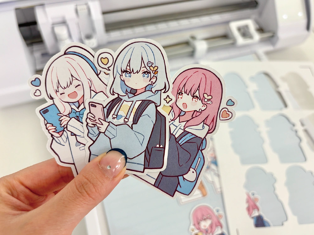手に持たれたスマートフォンを持つアニメ風キャラクターのステッカー3枚。背景にはカッティングマシンとカット中のステッカーが見える。