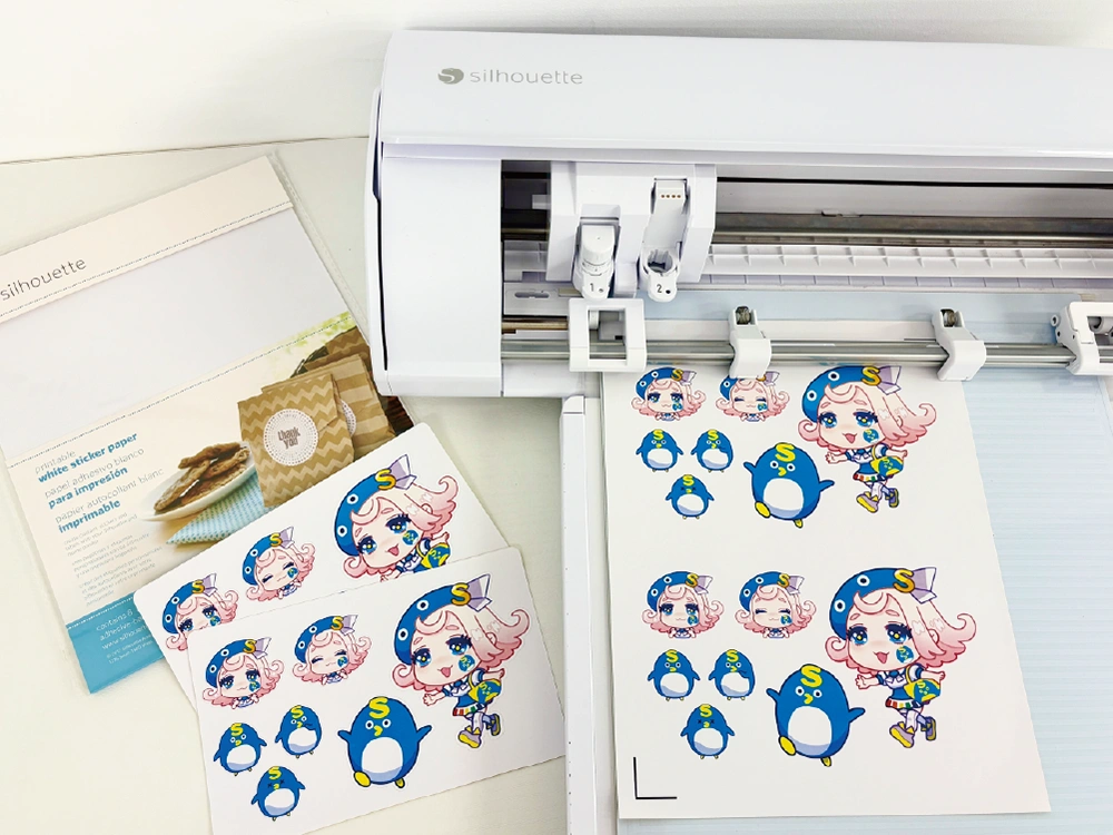カッティングマシンでアニメキャラクターとペンギンのステッカーがカットされている様子。左側には冊子も置かれている。