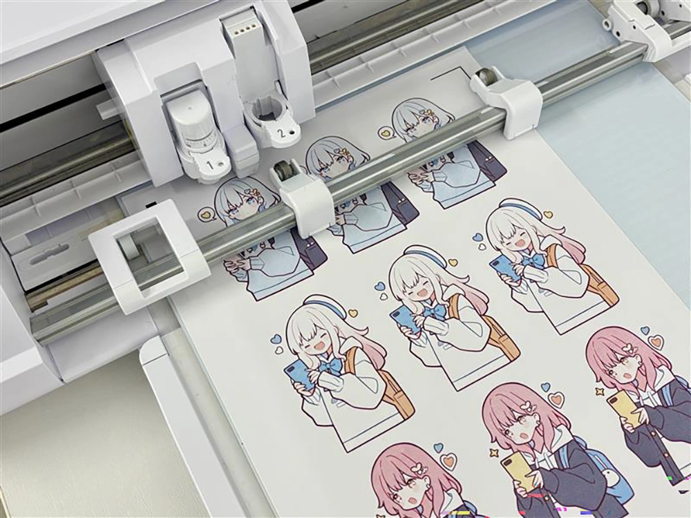 カッティングマシンが、スマートフォンを持ったアニメ風キャラクターのステッカーを複数枚カットしている場面。