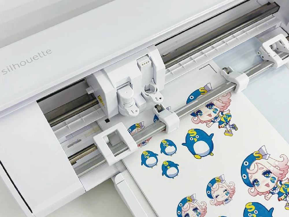 Silhouetteカッティングマシンが、女の子とペンギンのキャラクターのステッカーをカットしている様子。